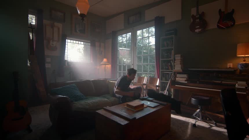 The video shows a cozy living room with a man relaxing on a couch surrounded by musical instruments and bookshelves a dog walks past in the foreground then jumps onto the couch to join the man who then pets it