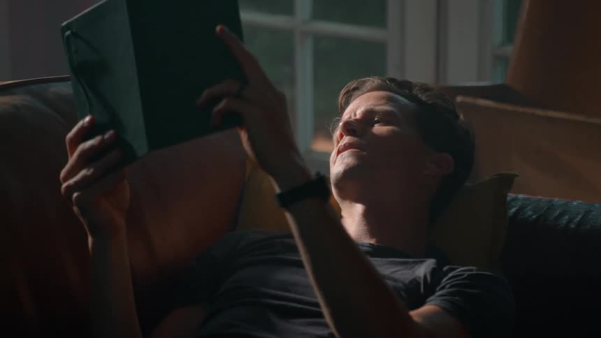 A man reclines on a couch reading a book bathed in soft natural light through nearby windows