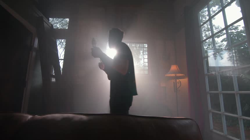 Light pours through a window behind a person playing guitar in a dim room with a lamp casting warm light across nearby furniture