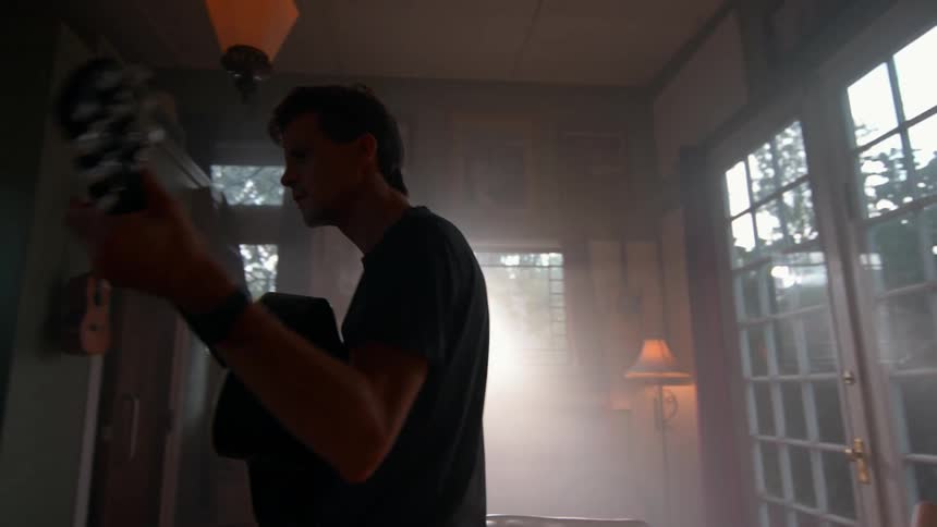 A man plays guitar in a sunlit room with bookshelves and large windows