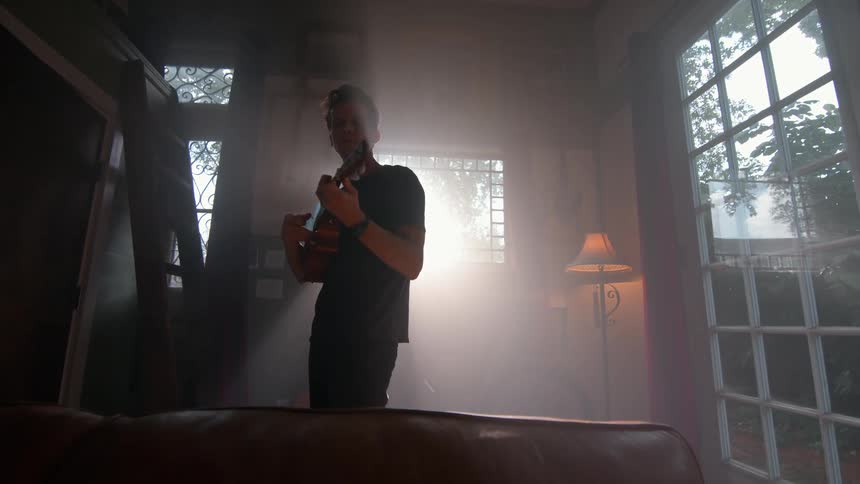 Young man plays a guitar while standing in a living room during daytime