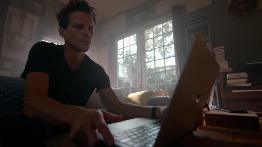 A person sits on a couch using a laptop in a cozy bookfilled room with large windows letting in natural light