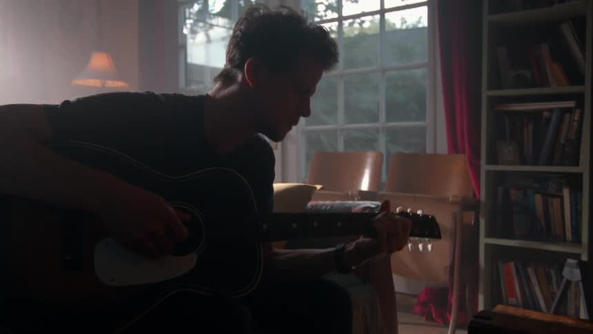 A musician sits in a sunlit room playing an acoustic guitar the room is decorated with various musical instruments and bookshelves filled with books
