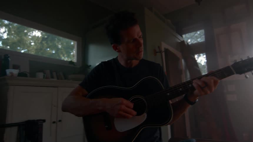 Male musician plays a guitar while standing in a living room during daytime