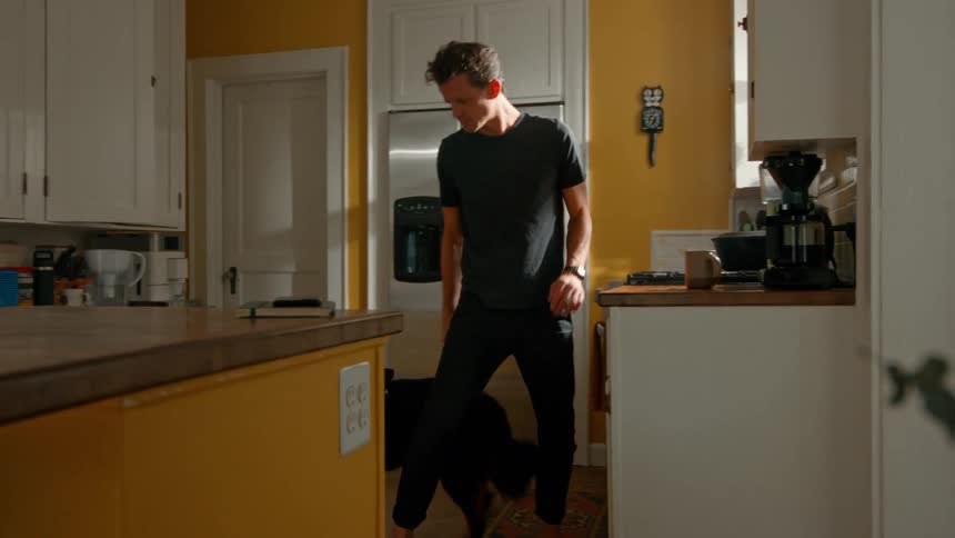 The video features a man in a black tshirt and pants in a brightly lit kitchen with yellow walls and white cabinets he is seen petting a black dog and the scene includes a refrigerator stove and coffee maker
