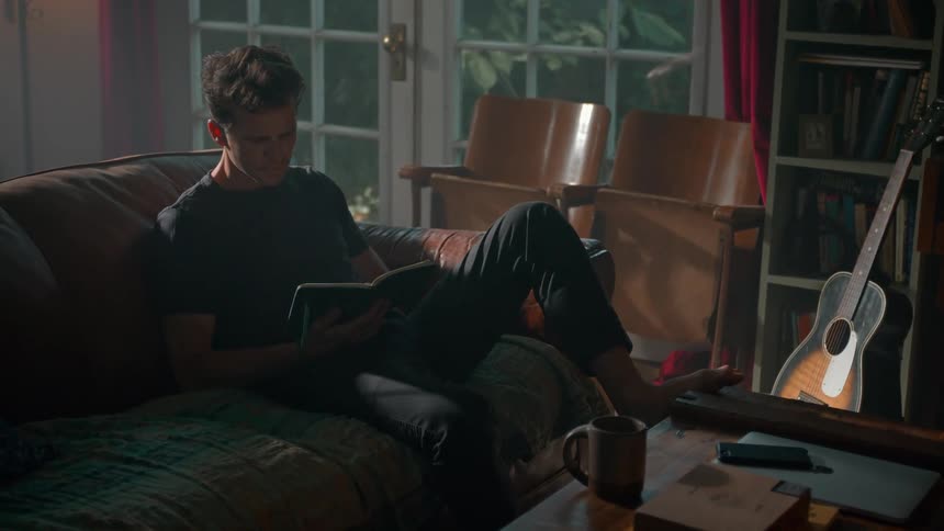 A person sits on a couch reading a book beside a table with a guitar coffee mug and notebook in a warmly lit room with a bookshelf and chairs in the background