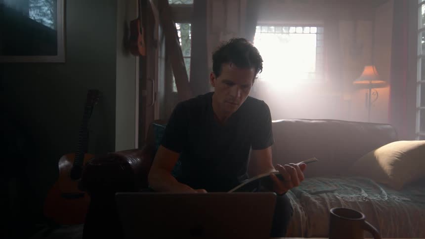 A young man is sitting on a couch in his living room at midday reading a book and typing on a laptop in front of him