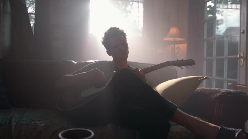 A man plays guitar on a sofa as sunlight streams through the window casting a soft glow