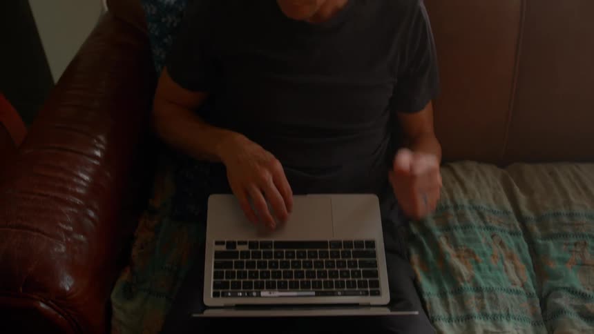 A man sitting on the sofa using a laptop and reaching into his hair to scratch his head