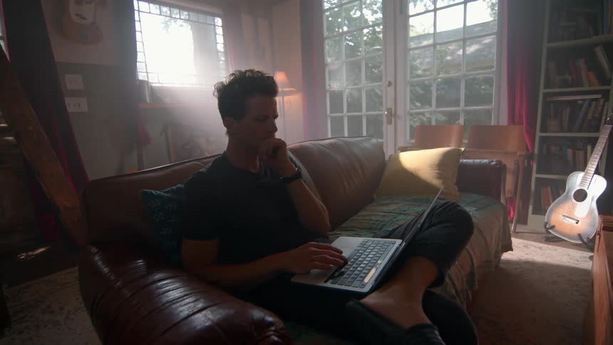 A person sits comfortably on a brown leather sofa engrossed in a laptop the room is softly lit by natural light streaming through large windows creating a peaceful atmosphere