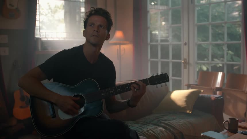 Man plays guitar while sitting on a bed in a softly lit room
