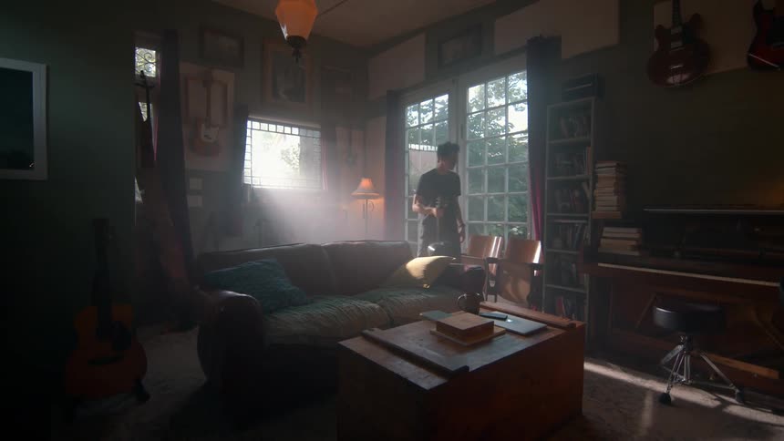 A person plays an acoustic guitar near a sunlit window in a cozy room filled with a piano guitars bookshelves and a brown couch beside a wooden table