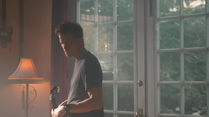 Male strumming a guitar then angrily setting it near a bookshelf