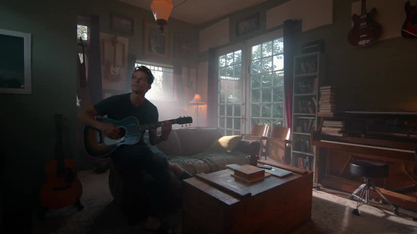A musician sits on a sofa playing an acoustic guitar in a room filled with musical instruments and books sunlight streams in through large windows illuminating the space