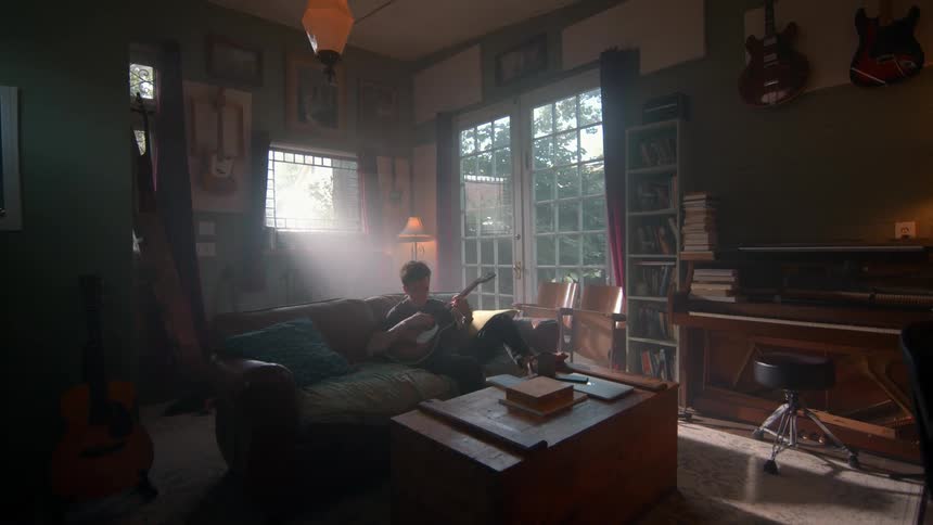 A man stands in a cozy musicfilled living room holding a guitar surrounded by instruments and books