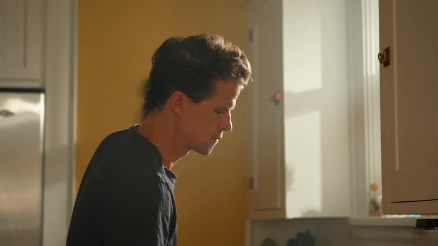 A man stands in the kitchen staring out the window as he serves coffee into a mug then leans against the countertop to relax and drink it