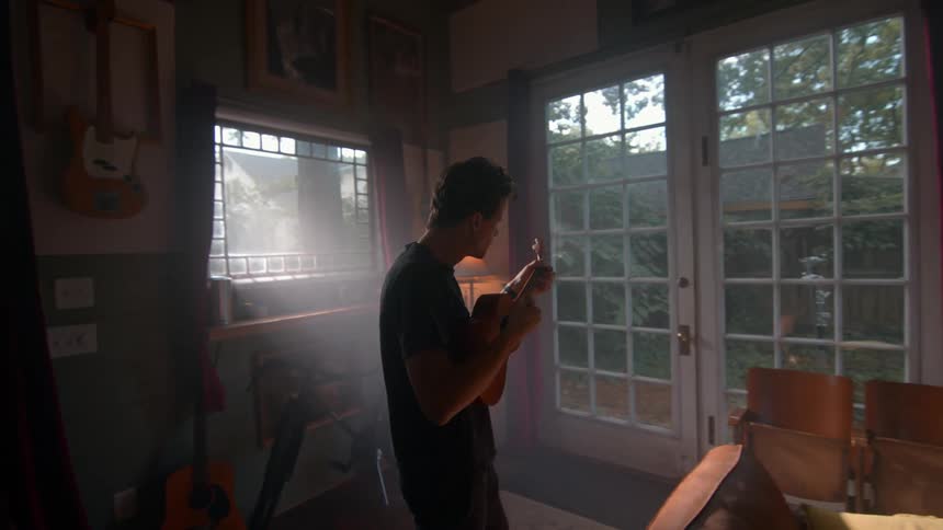 A male musician picks up a small stringed instrument from a wall and starts playing it in a living room during daytime