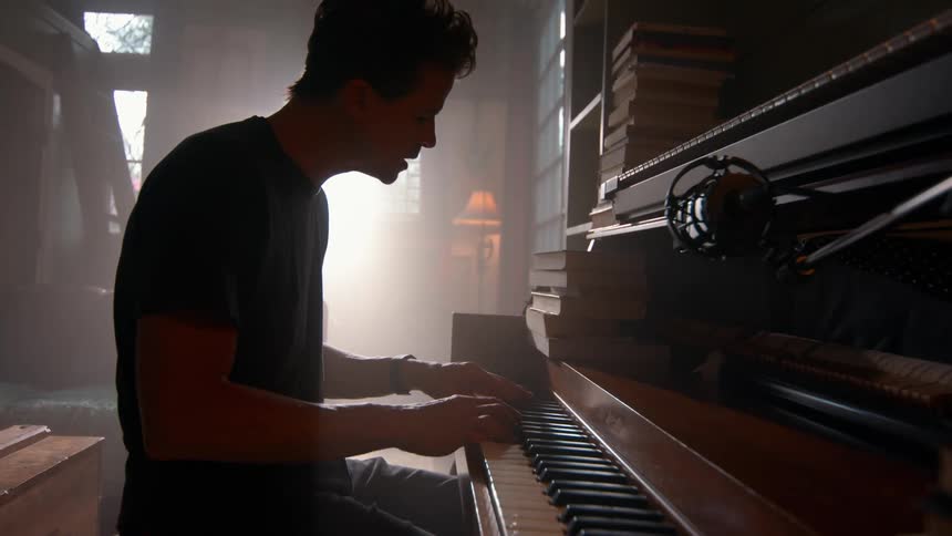 A man plays a piano in a warmly lit room filled with books deeply focused on his music