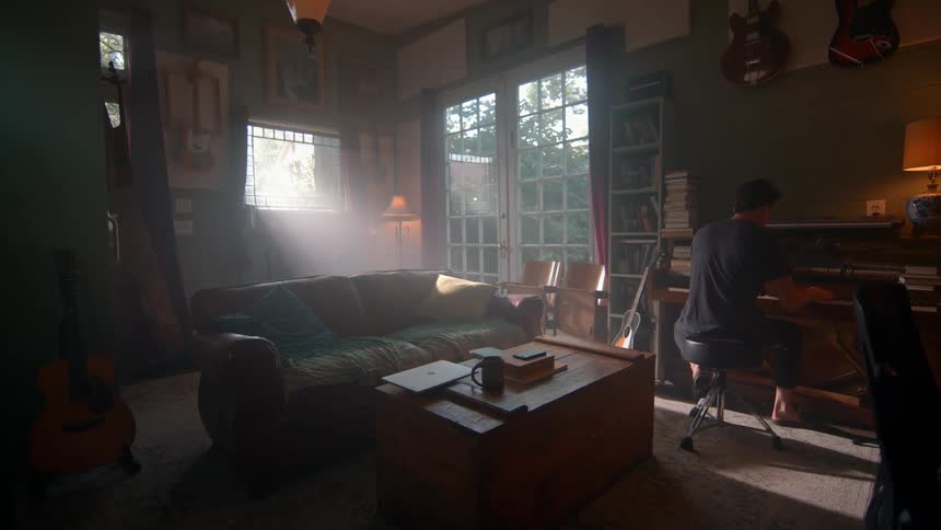 A person plays piano in a sunlit room with green walls surrounded by guitars bookshelves and a brown leather couch