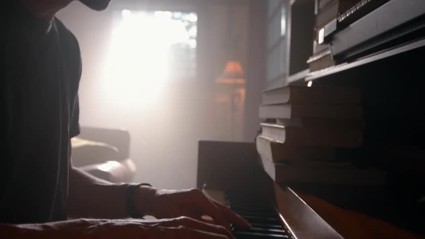 The silhouette of a man sitting playing the piano and there is strong natural light coming from the window