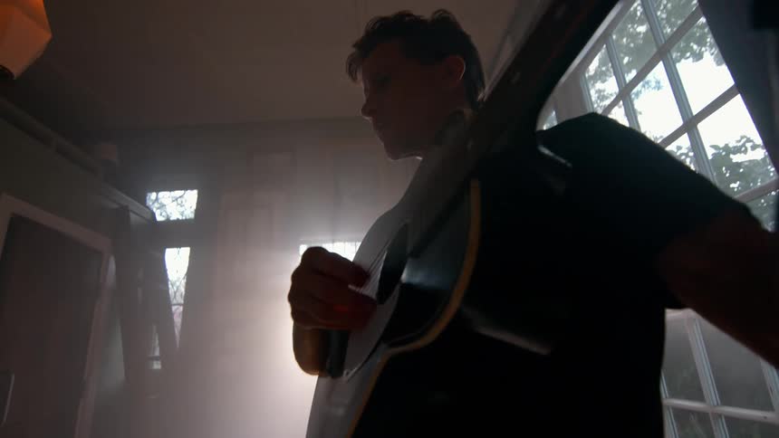 A young man is playing his guitar in his living room at midday