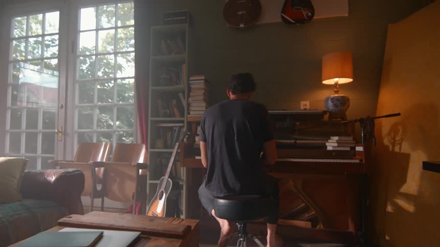 A man plays the piano in a sunlit room with green walls guitars bookshelves and a brown leather couch