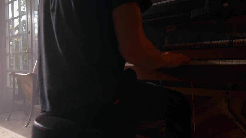 A barefoot man plays the piano in a dimly lit cozy living room beside tall windows glowing with morning light