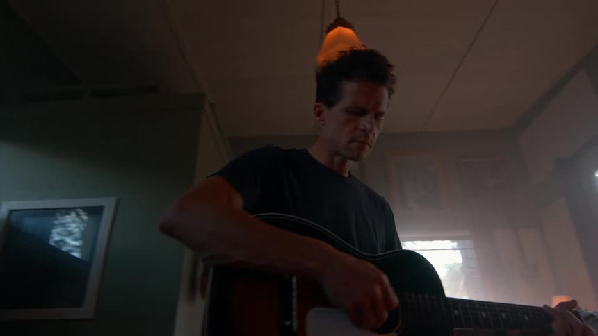 A person plays acoustic guitar in a dim room with hanging lights facing a window partially covered by blinds