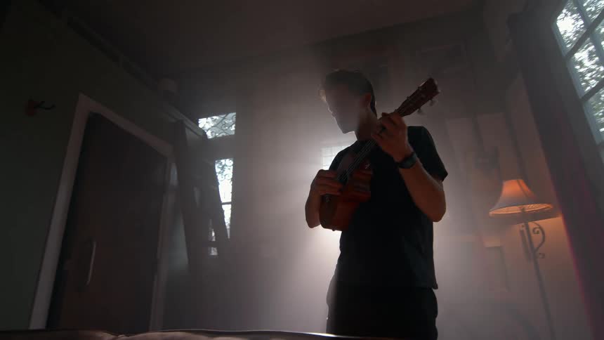 A man plays a ukulele in a dim hazy room with sunlight streaming dramatically through the window