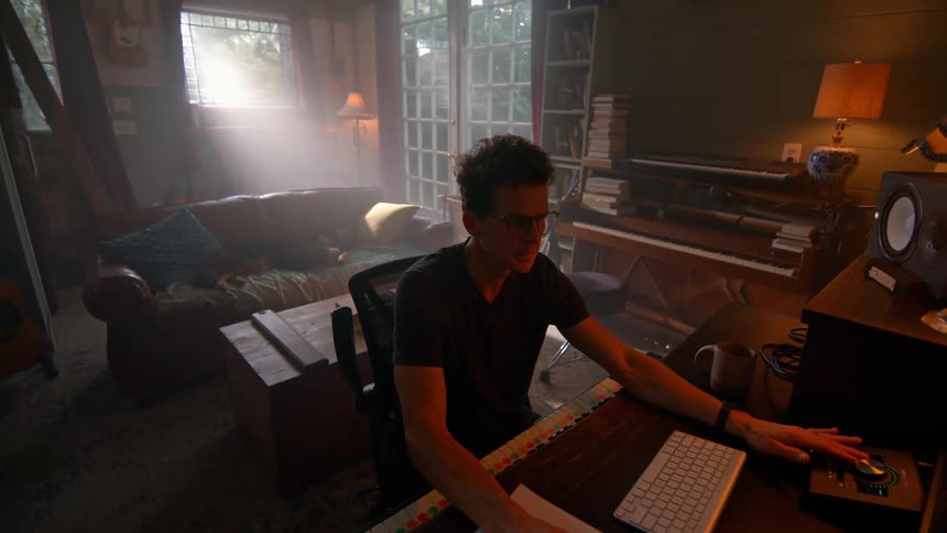 Young male musician sits at a desk in a living room while working with a desktop computer and musical equipment
