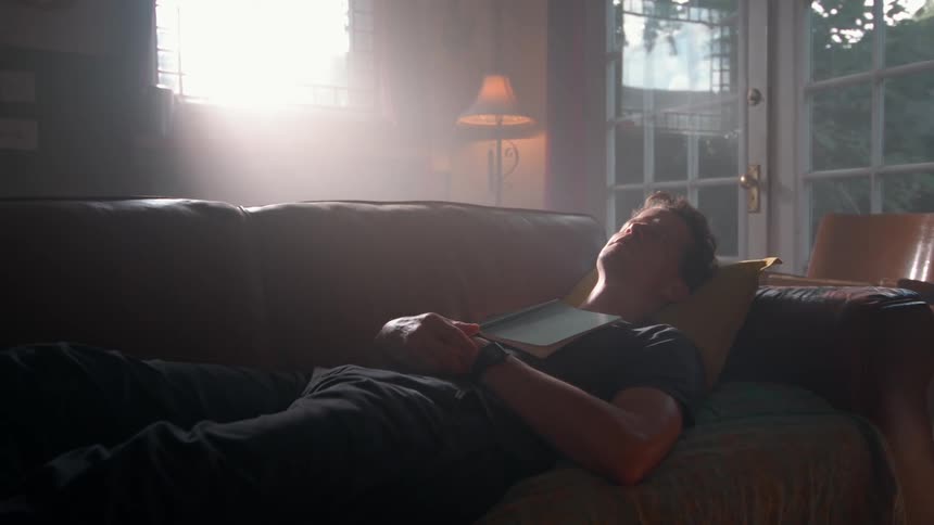 Man takes a nap on the couch with a book open on his chest while bright sunlight shines through the window