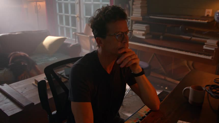 A person sits at a desk in a room with a piano bookshelves a coffee mug and a dog lying on the floor nearby