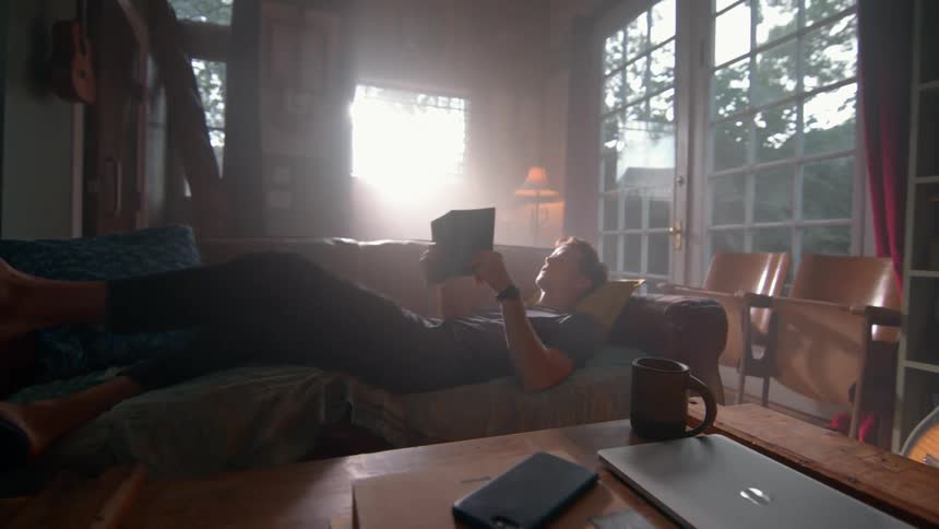 A person lounges on a sofa reading a book in a cozy sunlit living room