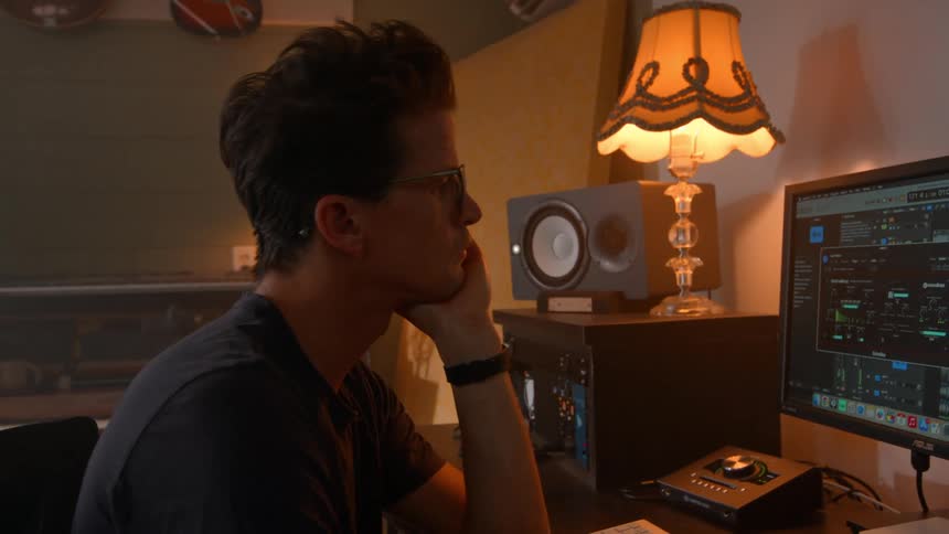 A person works at a desk with audio editing software on a monitor surrounded by sound equipment and lit by a desk lamp