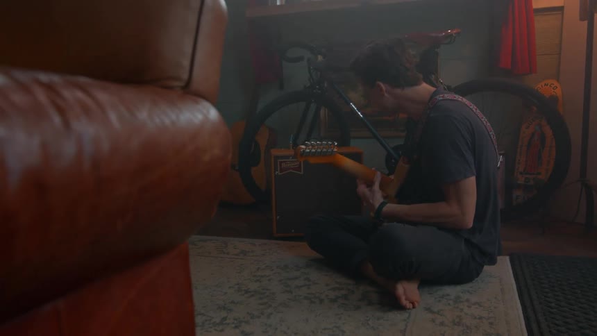 A person sits on the floor playing an electric guitar beside a small amplifier with a bicycle leaning against the wall and a leather chair nearby