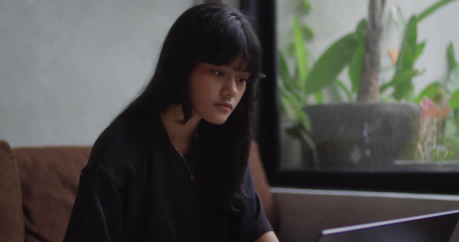A young girl sitting on a sofa next to a window and using a laptop