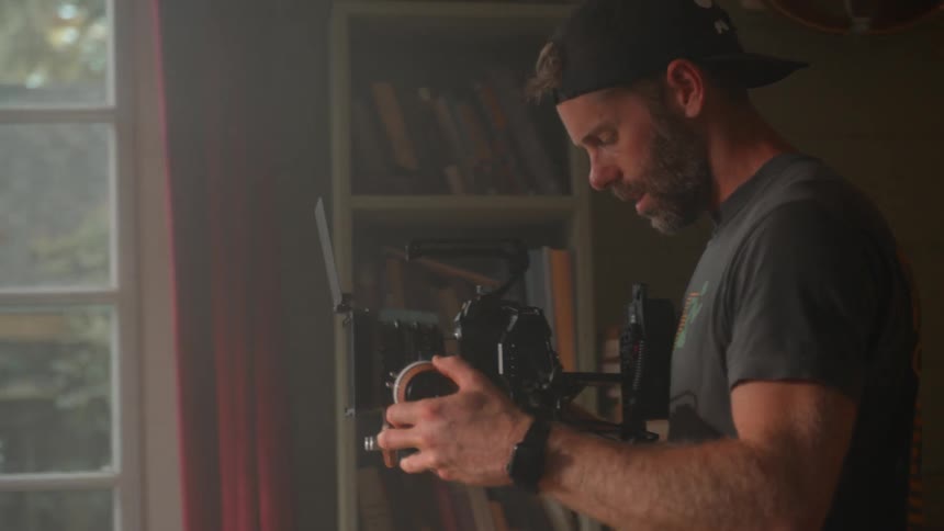 The video shows a man with a beard and a cap holding a camera rig adjusting settings and looking intently at the equipment he is standing indoors with a bookshelf visible on one side and a window in the background suggesting he is preparing for shoot