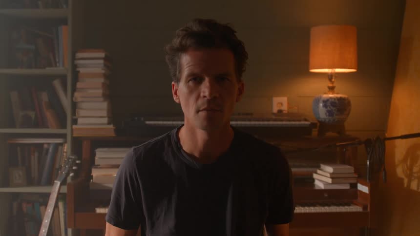 The video features a man with dark hair wearing a black tshirt standing in a dimly lit room with bookshelves and musical instruments in the background he looks directly at the camera with a neutral expression