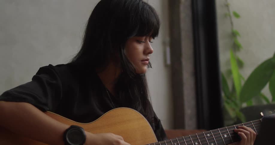 A young woman plays an acoustic guitar by a window absorbed in her music