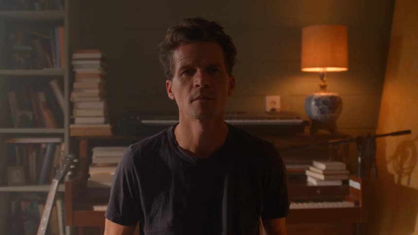 A person stands in a room surrounded by a guitar a piano stacked books and a small table with a lamp