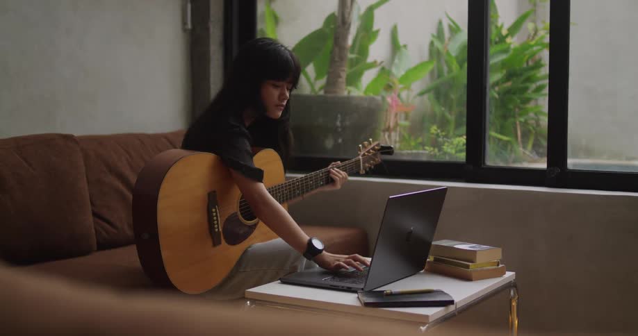 The girl is typing on a laptop while holding a guitar