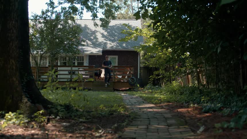 A person and a dog walk along a brick path from a house flanked by dense trees and overgrown foliage