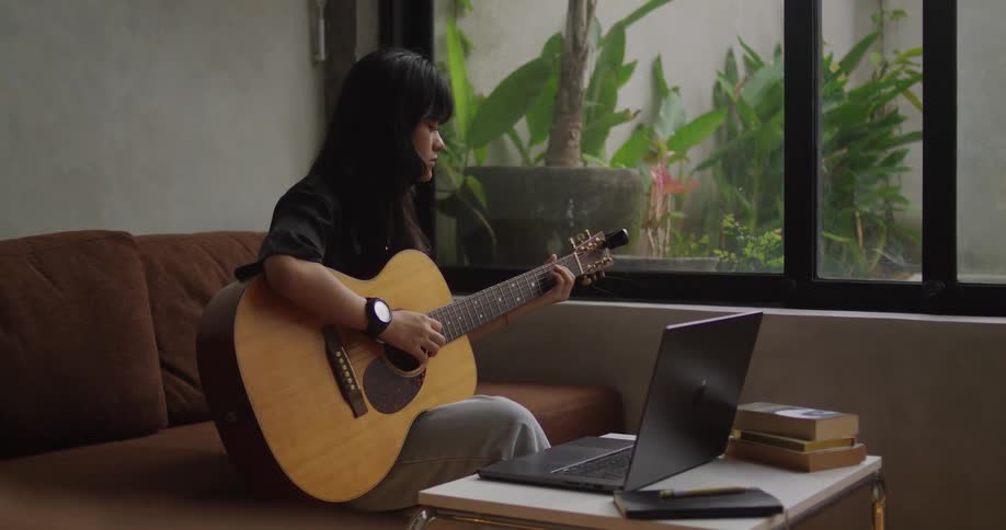 A young woman playing a guitar while sitting on a sofa in front of a laptop