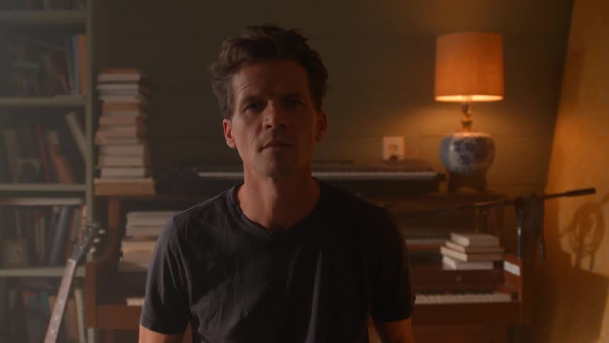 A person stands in a cozy room surrounded by books a guitar and a piano with warm light from a lamp in the background