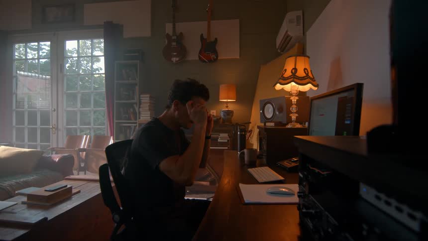 A person works at a desk with audio editing software on a monitor surrounded by sound equipment and lit by a desk lamp