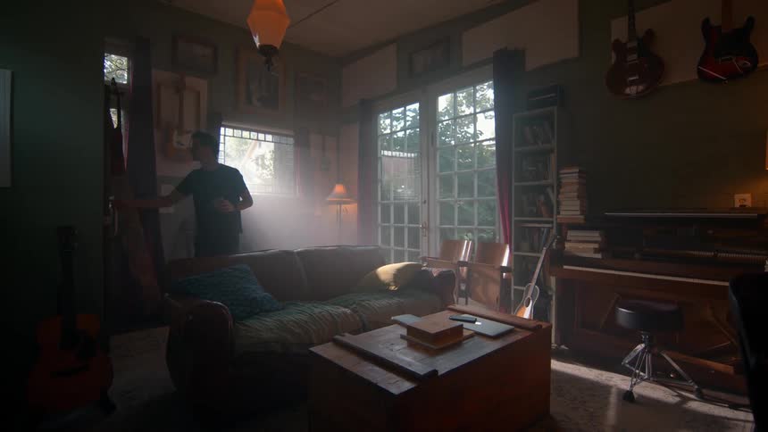 A man walks out of a sunlit room with dark green walls a brown leather couch and several musical instruments