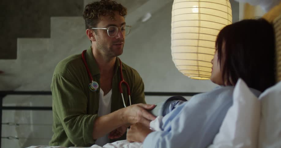 A person in a green jacket with a stethoscope places a blood pressure cuff on the arm of someone lying in bed next a large overhead lamp