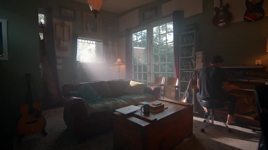 The video shows a man with dark hair wearing a black tshirt sitting at a piano in a room filled with musical instruments and bookshelves sunlight streams through a large window illuminating the space as he plays the piano