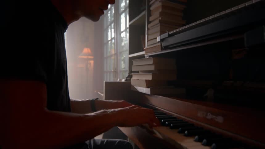 A young man is playing the piano in the living room where shelves are filled with books its midday and he is singing along enjoying the moment