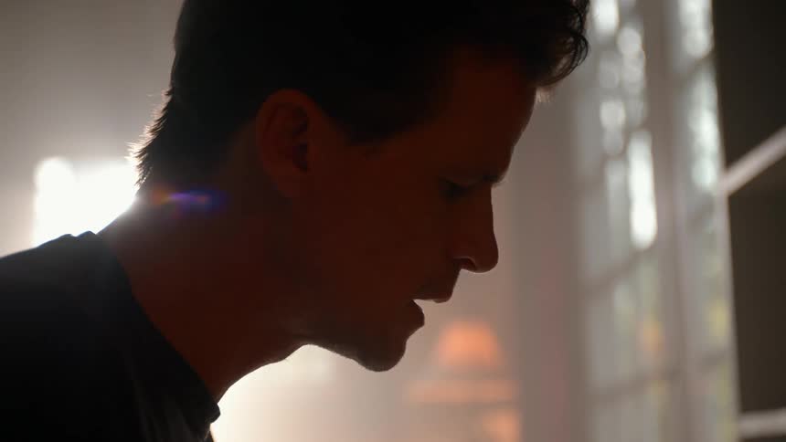 A person stands in a sunlit room near a window with sheer curtains playing piano